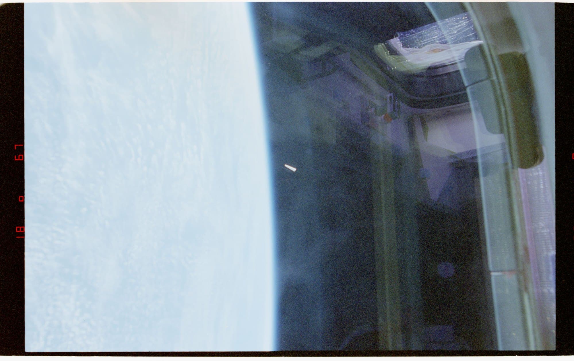 NASA-UAP-D030 — a photograph taken from Space Shuttle Columbia during STS-80 in 1996. The cloud-covered limb of the Earth curves across the left of the frame; the orbiter's window frame and payload-bay structure fill the right. A small, bright, elongated white object is visible in the dark space between them. Released by the Department of War in PURSUE Release 04.
